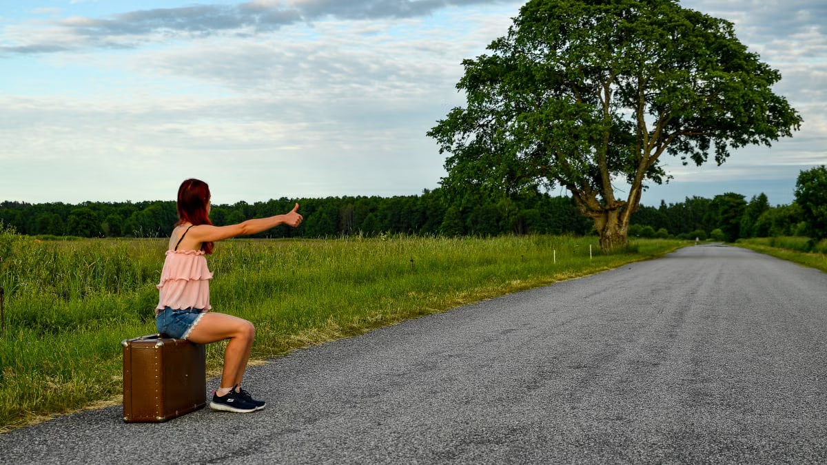 A woman sits on a suitcase, hitchhiking along a peaceful country road in Kolga, Estonia.