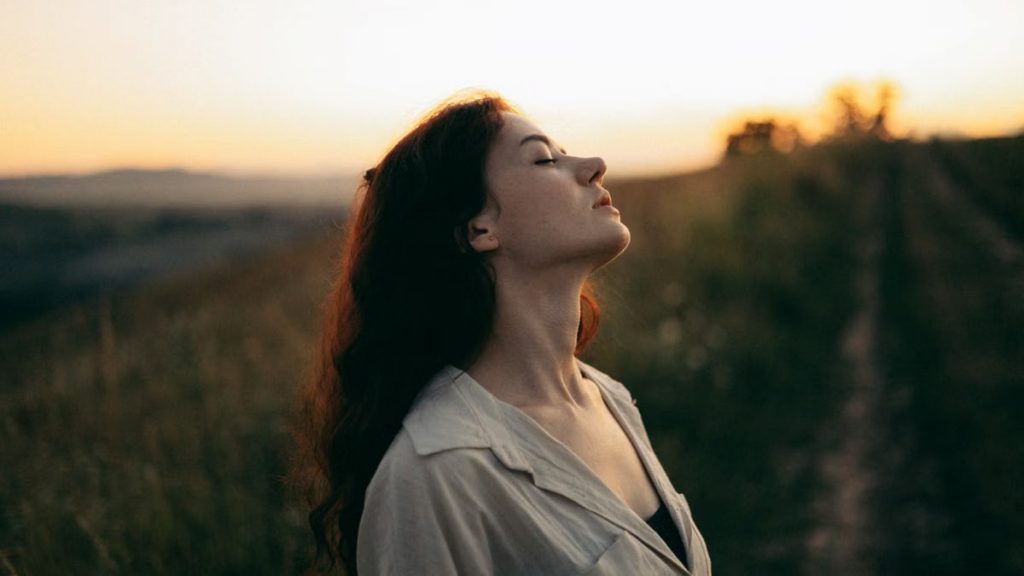 Woman standing with eyes closed in a field during sunset, exuding tranquility and peace.