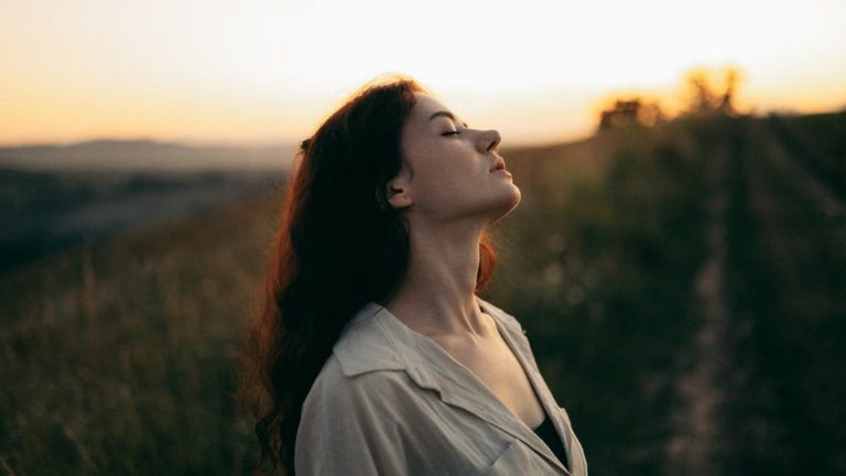 Woman standing with eyes closed in a field during sunset, exuding tranquility and peace.